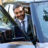 A smiling middle-aged man in a sharp business suit steps out of a modern car, looking friendly.