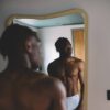 tobi-thomas-13 A fit shirtless man looks into a mirror in a bright modern bedroom, reflecting his toned physique.