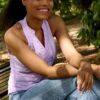 A smiling model with braided hair sits on a wooden park bench wearing a light purple top.