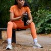 A fit model in orange activewear sits on a park bench holding a black water bottle outdoors.