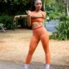A fit model in orange activewear performs a warm-up stretch in a green, leafy public park setting.