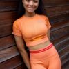 A smiling fitness model wearing an orange workout set poses against a wooden wall in bright sunlight.