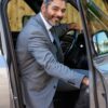 A smiling middle-aged man in a sharp grey suit sits in a luxury car, radiating professional warmth.