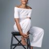 A graceful mature woman with a warm smile poses sitting on a black stool against grey backdrop.