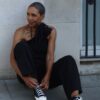 A stylish mature woman with short grey hair ties her sneakers while sitting against a stone wall.
