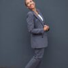 A smiling mature woman in a grey suit and white trainers stands confidently against a dark wall.