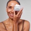 A smiling mature woman holds a jar of Medik8 skincare cream in front of her eye.