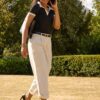A smiling young woman in business casual attire walks through a sunny park while talking on the phone.