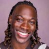 tobi-thomas-1 A handsome man with braided hair wearing an argyle sweater vest smiles warmly in a studio headshot.