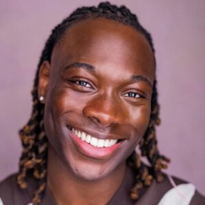 A handsome man with braided hair wearing an argyle sweater vest smiles warmly in a studio headshot.