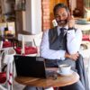 A smiling middle-aged man in a suit vest sits at a café table with his laptop.