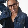 A stylish mature woman with short grey hair wears glasses and a denim jacket, smiling warmly outdoors.