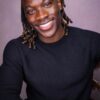 tobi-thomas-3 A handsome man with braided hair smiling brightly at the camera against a neutral studio background.