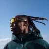 tobi-thomas-11 A man with dreadlocks wearing sports sunglasses and a technical jacket stands against a clear blue sky.