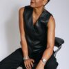 A mature woman with short dark hair wearing a black waistcoat sits elegantly on a studio stool.