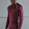 tobi-thomas-12 A fit male model wearing a maroon long-sleeved athletic top stands against a neutral grey studio background.