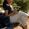A smiling young woman sits on a park bench working on a tablet in bright sunny weather.