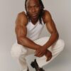 tobi-thomas-15 A muscular man with braided hair poses in a white tank top against a neutral studio background.