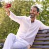 A smiling middle-aged man in a light linen shirt sits on a park bench taking a selfie.