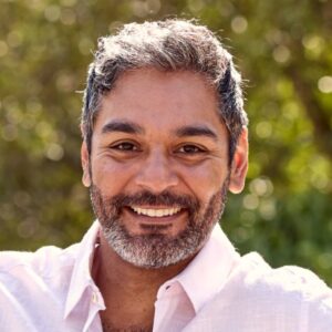 A smiling man with a grey beard sits on a park bench in bright, sunny, natural light.