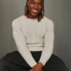 tobi-thomas-2 A handsome man with braided hair wearing a light sweater smiles warmly against a neutral grey background.