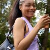A smiling young woman with braided hair holding a takeaway coffee cup in a sunny outdoor park setting.