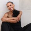 A smiling, mature woman with short grey hair sits against a neutral wall in a stylish outfit.