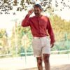 A stylish middle-aged man in a terracotta linen shirt walks outdoors through a sunlit park setting.