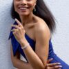 A smiling young woman in a blue dress with braided hair leans against a textured white wall.
