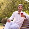 A smiling man sits on a sunny park bench, checking his smartphone in a relaxed, natural outdoor setting.