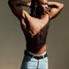 tobi-thomas-17 A muscular man with braided hair poses shirtless in dramatic side-lit shadows wearing light blue denim jeans.