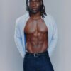 tobi-thomas-16 A muscular man with dreadlocks wears an open light blue shirt and dark jeans against a grey background.