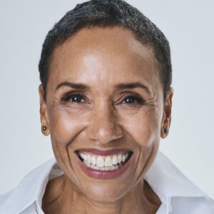 A warm, smiling mature woman with short hair wearing a crisp white shirt against a neutral background.