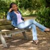 A smiling young woman in denim jacket and jeans poses on a wooden bench in a park.