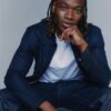 tobi-thomas-4 A stylish young man with braided hair sits against a neutral background wearing a casual navy blue jacket.