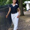 A smiling model with braided hair wearing smart casual clothing stands by a metal fence in a park.