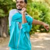 A smiling young man in a turquoise tracksuit stretches his arm outdoors in a sunny park setting.