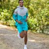 A smiling man wearing teal sportswear runs joyfully along a sunlit woodland path during the day.