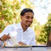 A smiling man in a white shirt works on a laptop in a bright, sunlit outdoor garden.