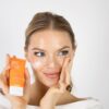 eloise-skinner-6 A smiling blonde woman applies orange-branded skincare moisturiser to her cheek against a clean white studio background.
