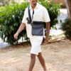 A smiling man wearing a white shirt and shorts walks through a sunlit park with a bag.