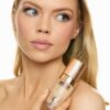 eloise-skinner-7 A blonde model posing with a bottle of luxury foundation, showcasing flawless skin in a bright studio setting.