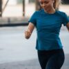 eloise-skinner-10 A fit young woman with a ponytail jogging outdoors, wearing blue sportswear and white wireless earbuds.