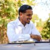 A smiling man in a white shirt works on his laptop at an outdoor table in sunshine.
