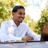 A smiling man in a white shirt works on his laptop at an outdoor table in nature.