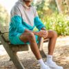 A smiling man in sportswear sits on a park bench holding a water bottle in bright sunlight.