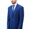 A man in a smart navy blue suit and tie stands against a plain white wall background.