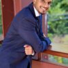 A smiling young man in a navy blue suit leans against a railing outdoors, looking professional yet approachable.