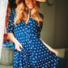 eloise-skinner-3 A smiling blonde woman wears a patterned blue summer dress and straw hat in a bright, indoor setting.