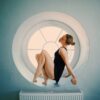 eloise-skinner-15 A woman in a black leotard sits gracefully in a large circular window, illuminated by soft natural light.